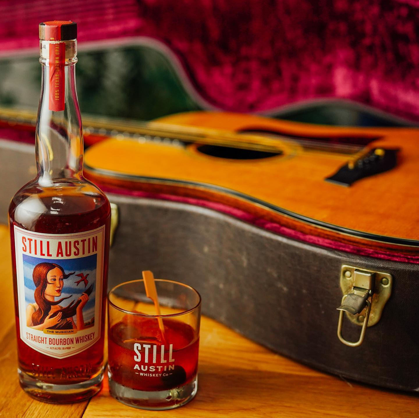 Still Austin Straight Bourbon "The Musician" sitting next to a still austin glass with whiseky. An acoustic guitar sits in case behind and out of focus